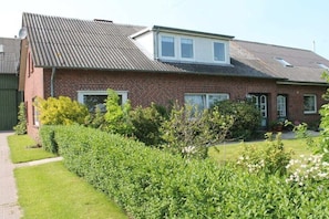 A two-story brick house with a sloped roof, a satellite dish, and a well-maintained garden with hedges and flowering plants. - Holiday apartment with 2 bedrooms (Midlum)