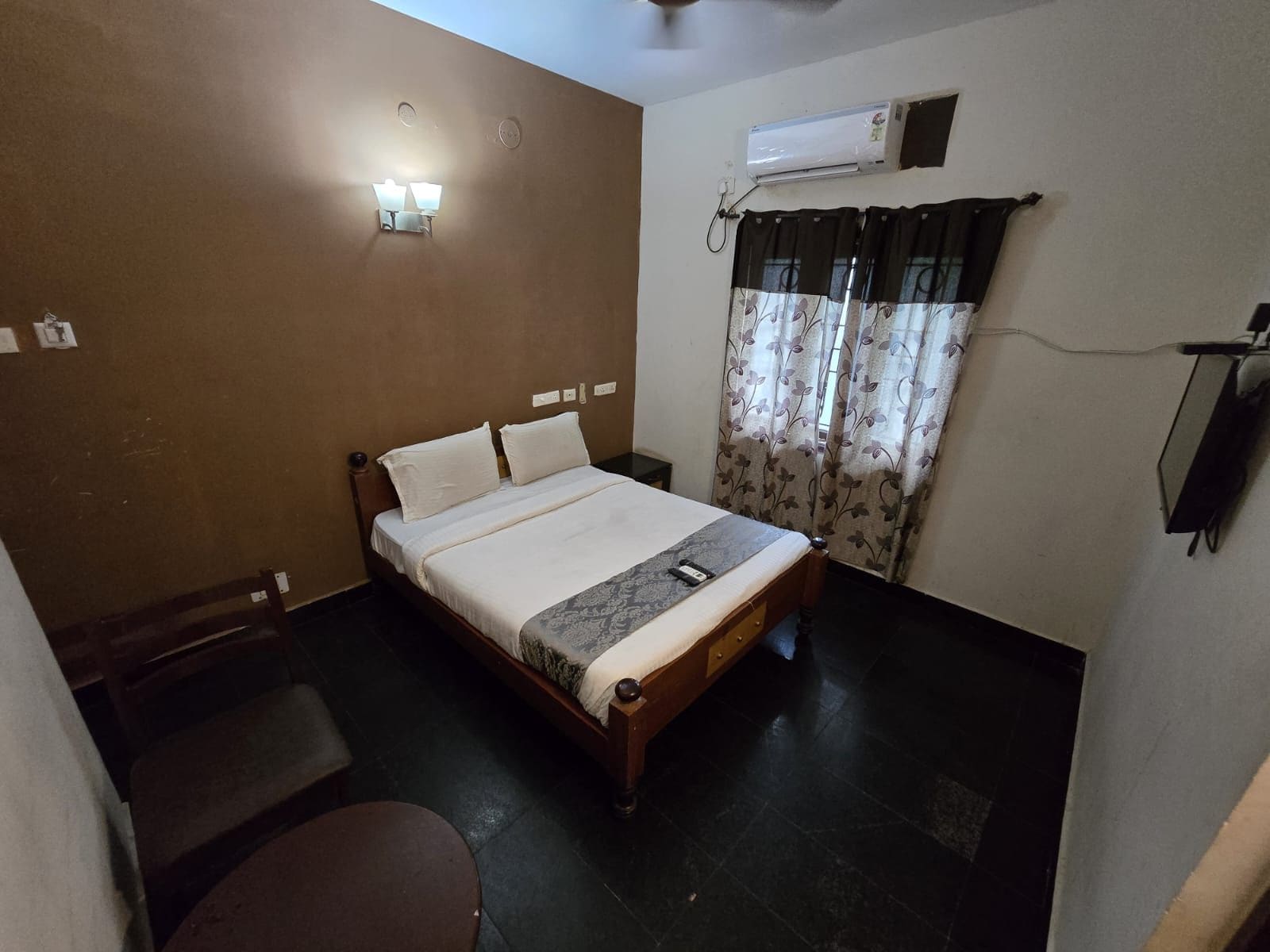 Un lit simple avec une tête de lit, une table de chevet, une chaise, un appareil de climatisation et une fenêtre avec des rideaux.