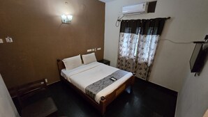 A single bed with a headboard, a bedside table, a chair, an air conditioning unit, and a window with curtains. - Grace Residency (MADURAI)