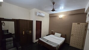 A hotel room with a bed, a desk, a chair, a wardrobe, and a door. - Grace Residency (MADURAI)