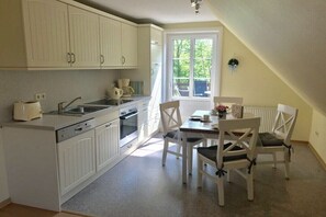 Et køkken med hvide skabe, et spisebord med stole og et vindue med udsigt til grøn vegetation.