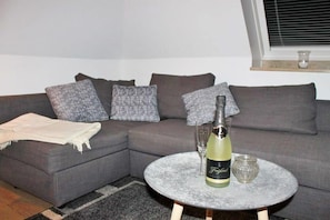 Ein Wohnzimmer mit einem grauen Ecksofa, einem runden Marmortisch, einer Champagnerflasche und zwei Gläsern.