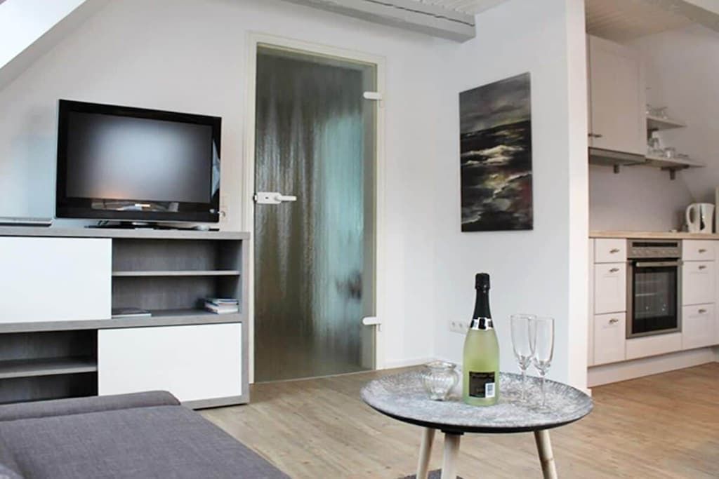 Una sala moderna con televisor de pantalla plana, una mesa de comedor con una botella y vasos, y una cocina al fondo.