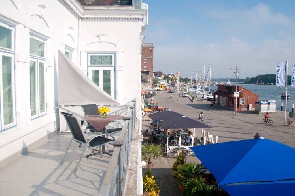 A balcony with outdoor seating, overlooking a marina with boats and a street with cyclists.