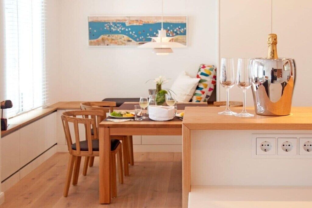 Un comedor con una mesa de madera, sillas, un jarrón blanco con flores y una botella de champán.