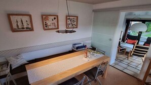 A dining area with a long wooden table, chairs, and a bench. There are framed artworks on the wall, a hanging light fixture, and a view through an open door. - Holiday home Seestern (Hasselberg)