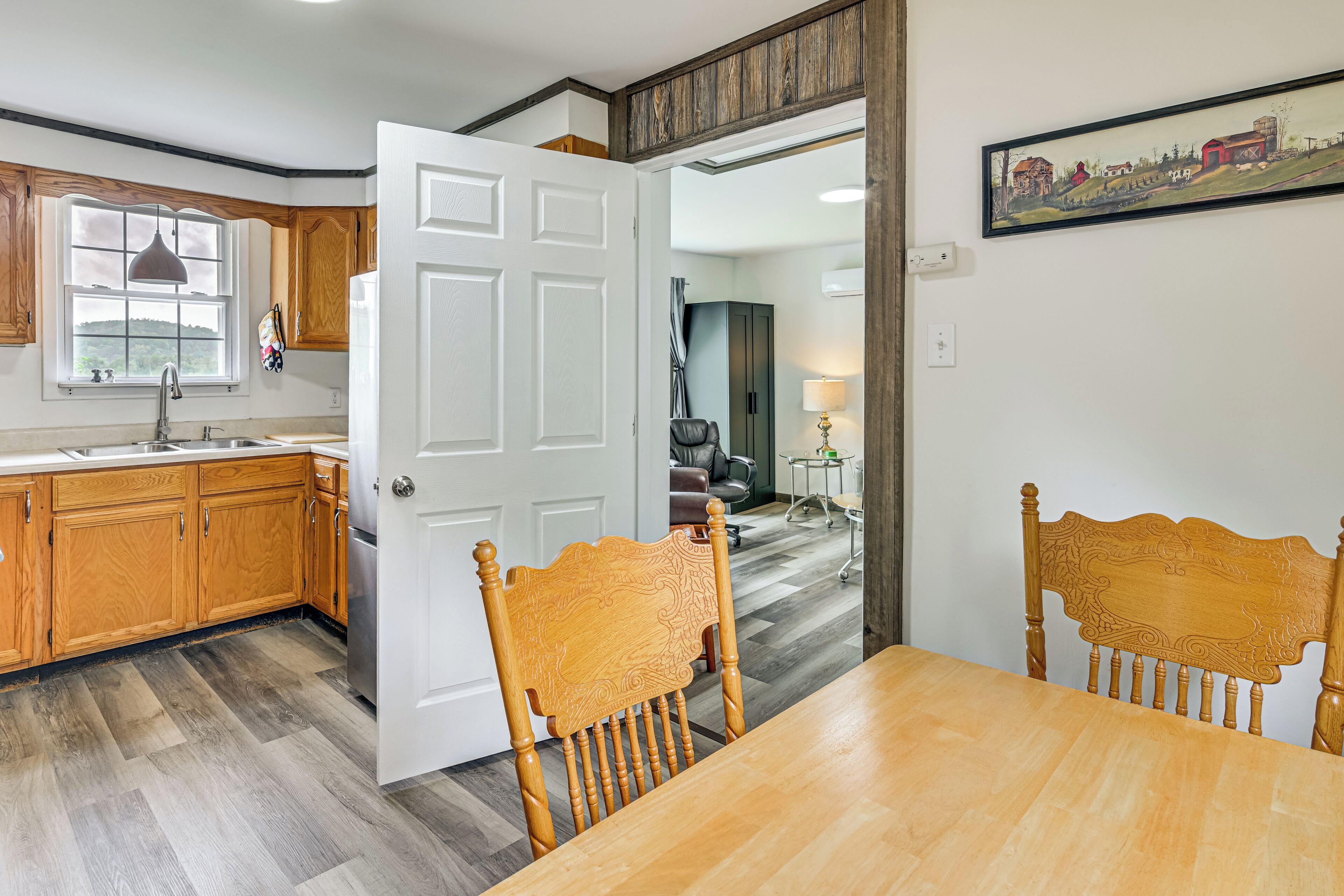 A modern kitchen with wooden cabinets, a dining area with a wooden table and chairs, and a view of a countryside landscape through a window.