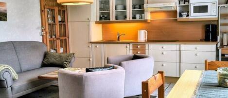 Una sala de estar moderna con un sofá, sillones, una mesa de centro y una mesa de comedor. Hay una cocina con armarios, un microondas y una cafetera.