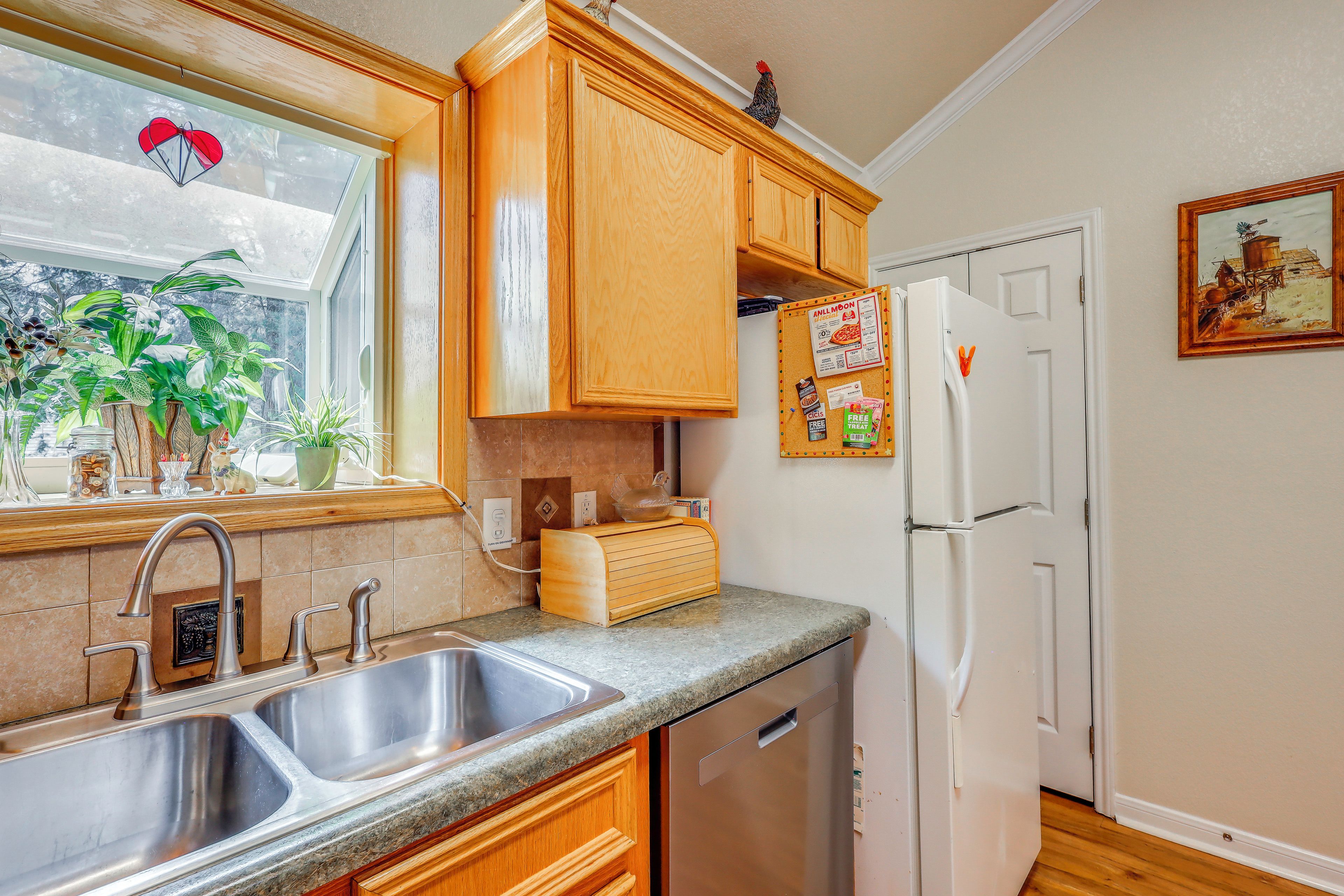 A kitchen with wooden cabinets, a stainless steel sink, a refrigerator, and a window with plants.