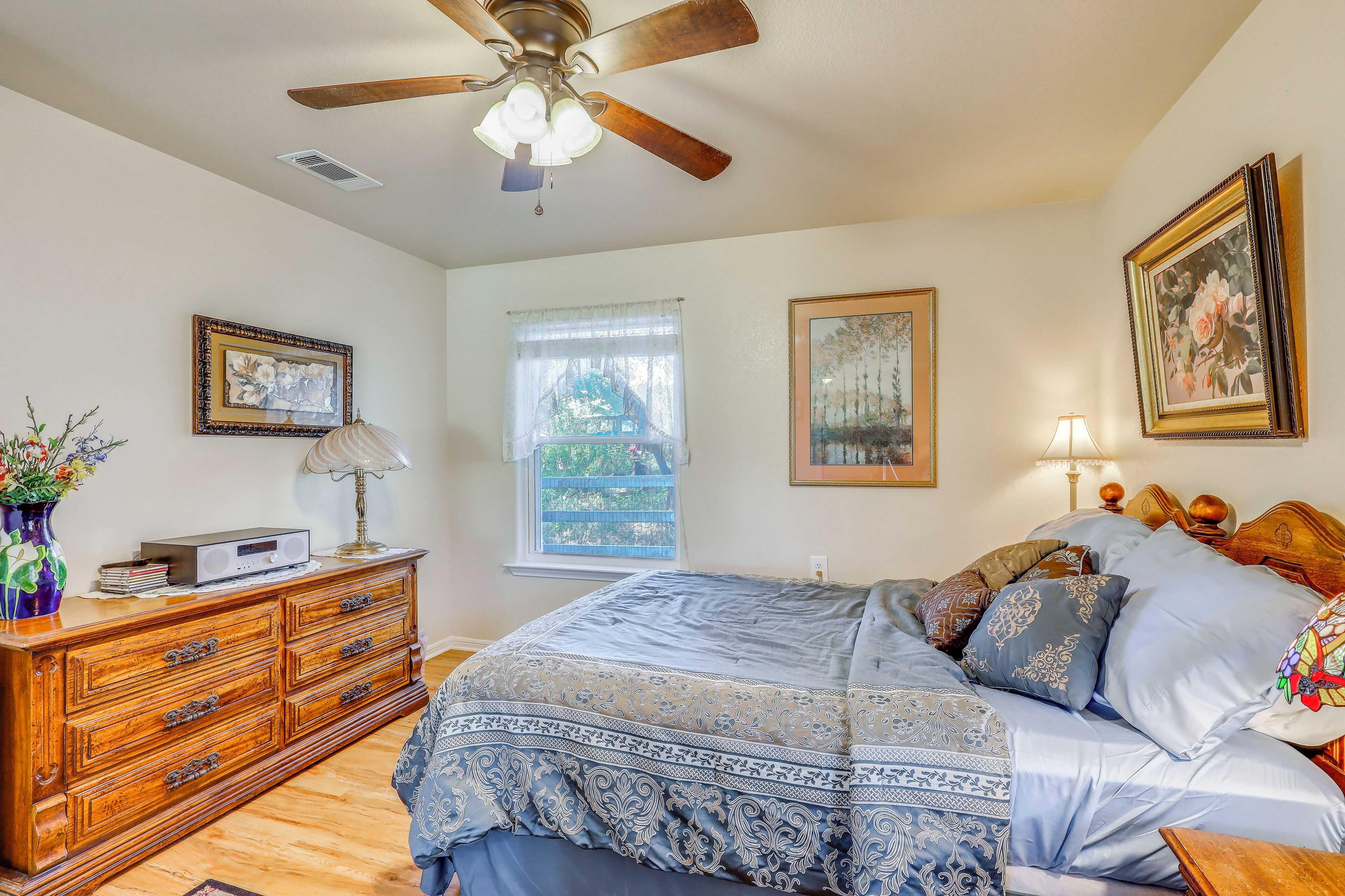 A bedroom with a bed, dresser, ceiling fan, window, and artwork.