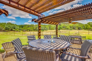 Een terras buiten met een ronde tafel en rieten stoelen, omgeven door een houten pergola.