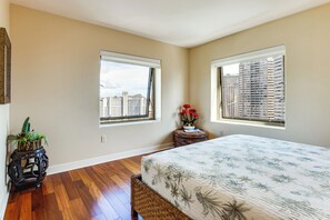 A bedroom with a large bed, two windows, a small side table with a vase of flowers, and a potted plant. - Walk to Beach, Shopping & Dining! Waikiki-Area (Honolulu)