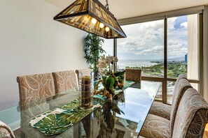 A dining area with a glass table, patterned cushions, a hanging plant, and a view of the ocean and cityscape through large windows. - Walk to Beach, Shopping & Dining! Waikiki-Area (Honolulu)