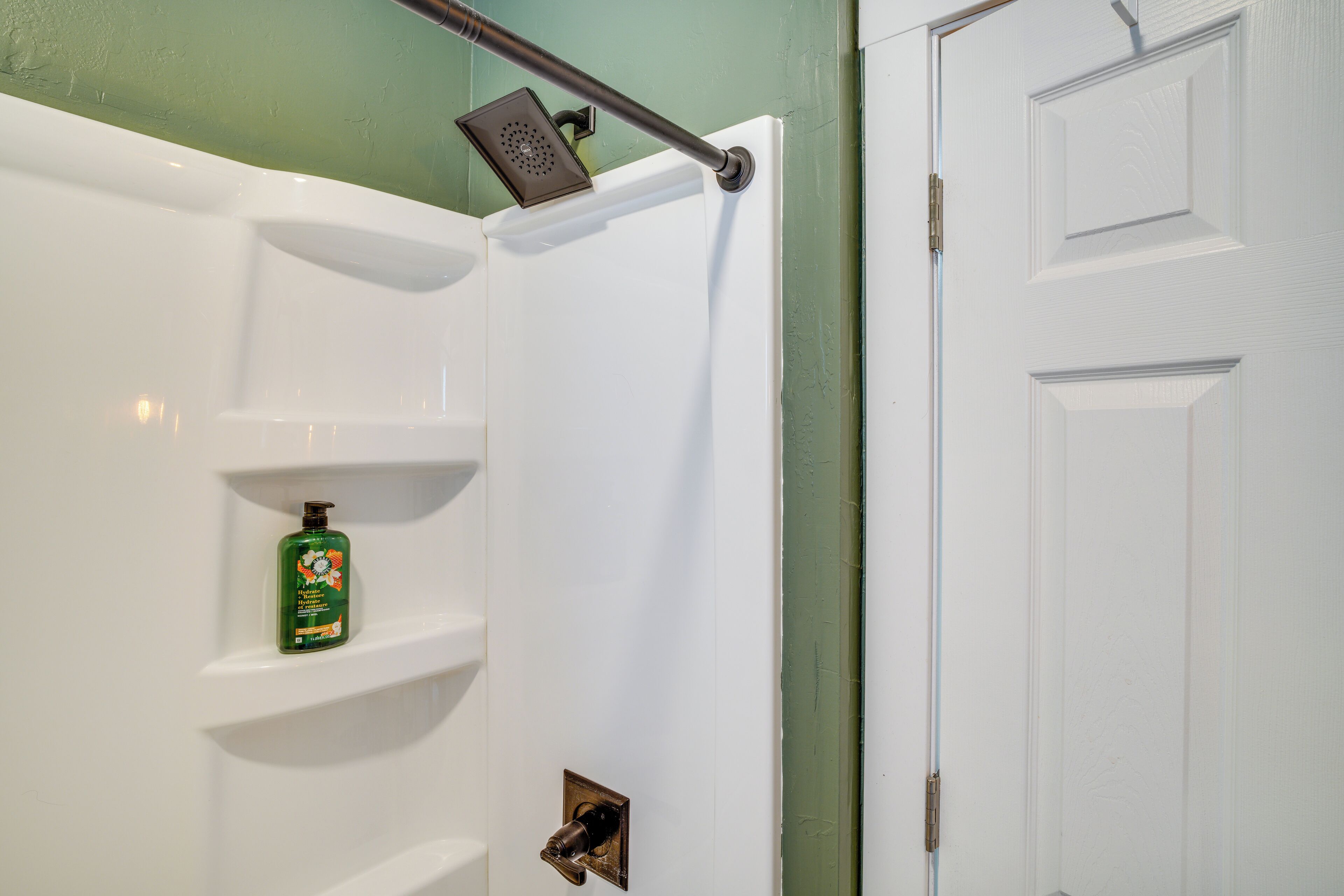 A bathroom with a shower, a white door, and a green bottle of shampoo on a shelf.