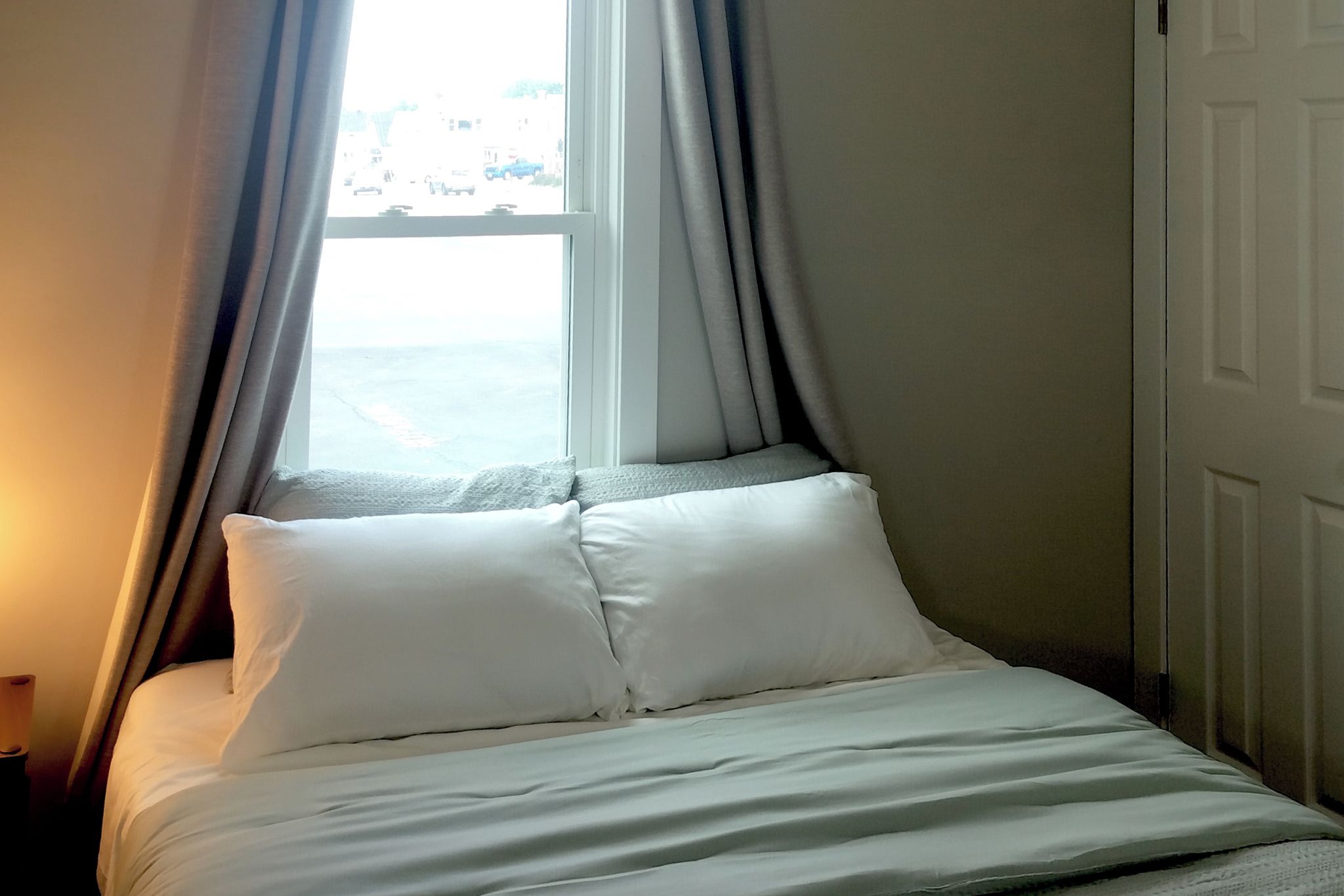 A bed with white bedding, a window with curtains, and a view of the street outside.
