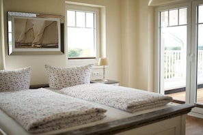 A bedroom with a bed, two pillows, a bedside table with a lamp, a framed picture on the wall, and a window with a view of the outdoors. - Villa Ocean Time WHG 17 (Haffkurg)