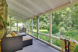 A covered outdoor seating area with a wicker sofa and a dining table, overlooking a lush garden. - Historic Streamside Farmhouse in Stroudsburg! (Stroudsburg)