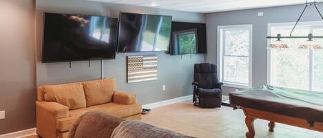 A living room with a sofa, a chair, a pool table, and two mounted televisions.