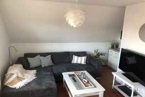 A modern living room with a sectional sofa, a coffee table with remote controls, a television on a stand, and a decorative hanging light fixture. - Holiday apartment in Upleward with balcony (Upleward)