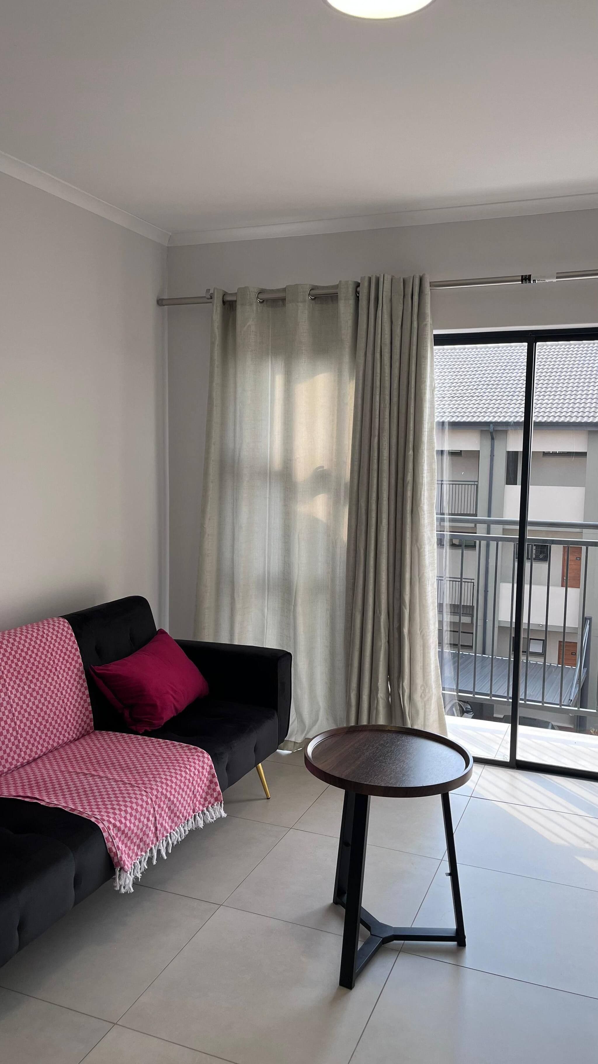 Una sala de estar moderna con un sofá negro, una mesa redonda y vistas a un edificio residencial a través de un ventanal amplio.