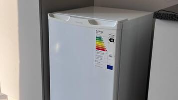Ein kompakter, weißer Hisense-Kühlschrank mit Energieeffizienzlabel steht in einer Ecke des Raumes, der mit Fliesen ausgelegt ist, und zur Linken ist eine Tür zu sehen.