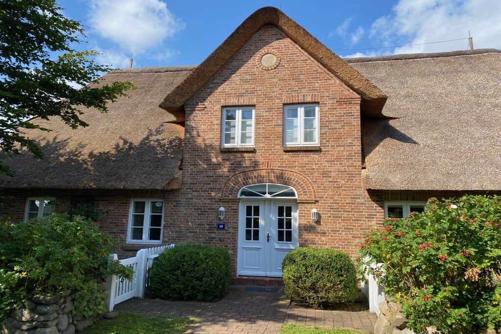 A brick house with a thatched roof, a white door, and a small garden with a white picket fence.