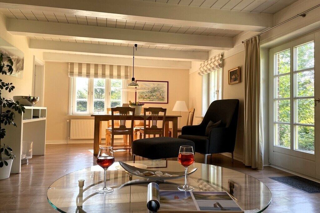 A living room with a glass coffee table, a dining area with wooden chairs, a black armchair, and a large window with curtains.