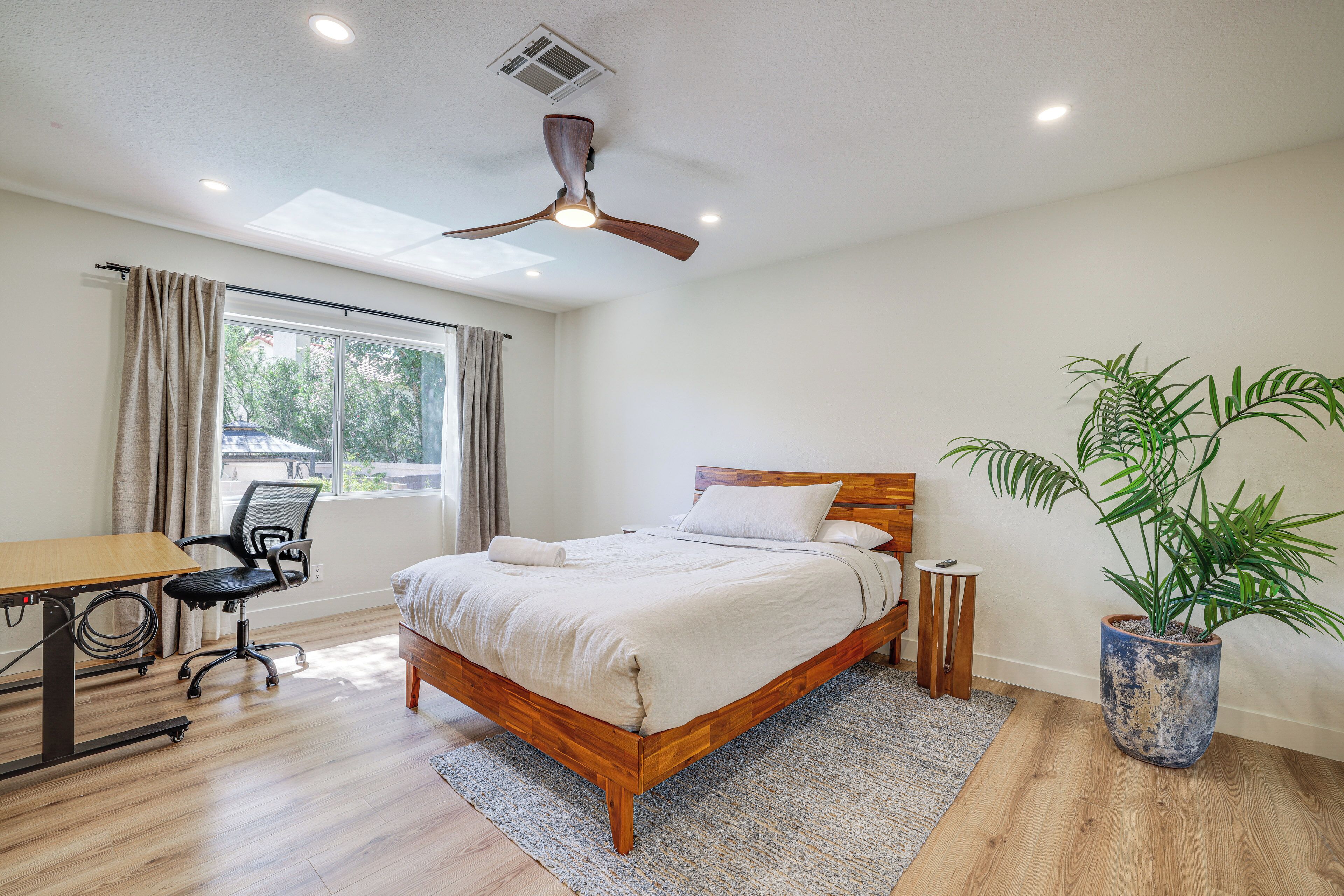 A bedroom with a bed, desk, chair, ceiling fan, window, and a potted plant.