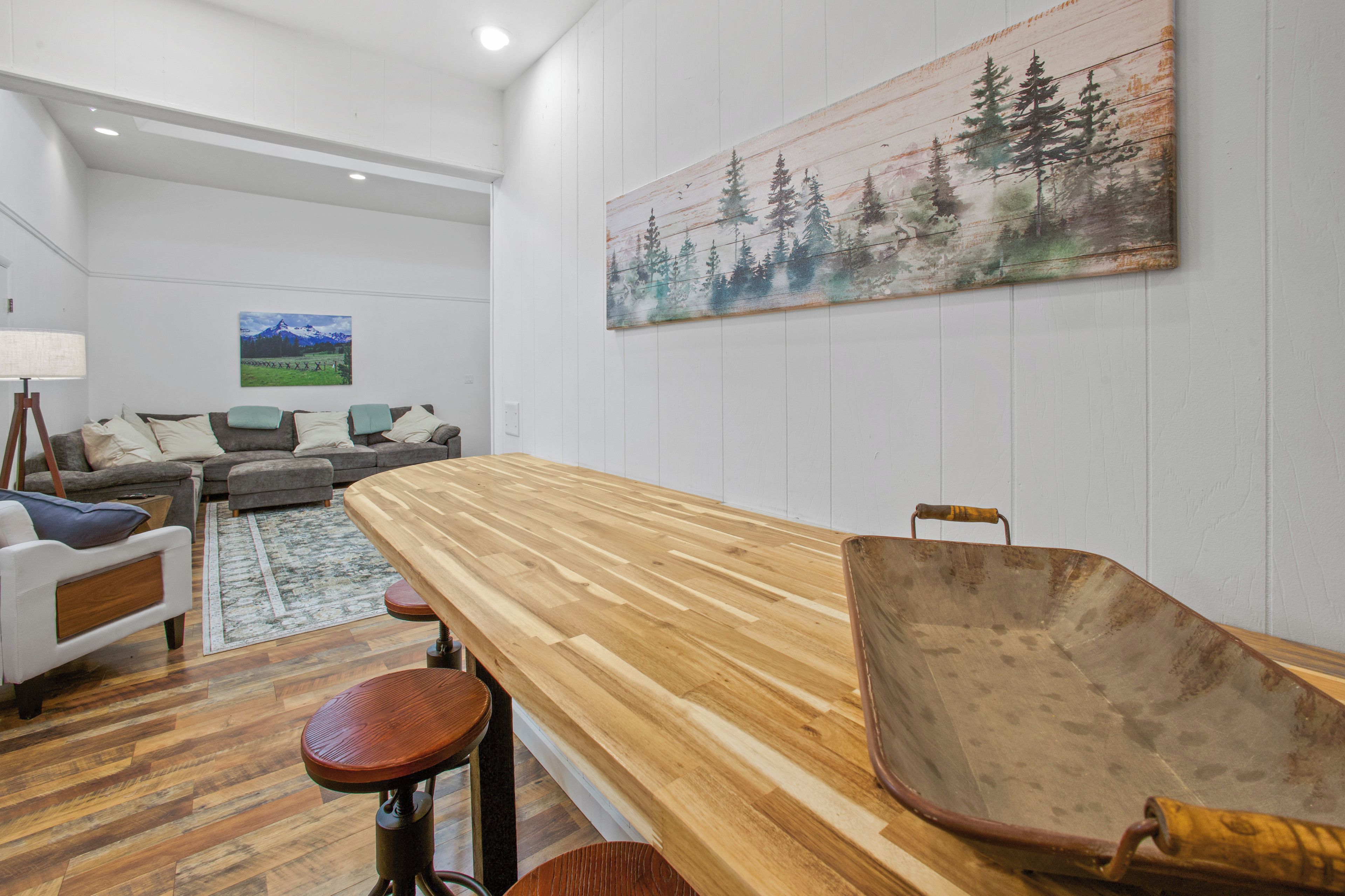 A modern living room with a wooden dining table, a wheelbarrow, and a painting of a forest on the wall.