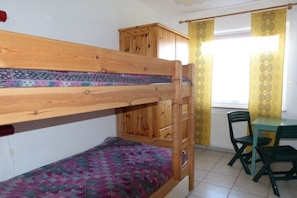 Una cama nido con colcha estampada, un armario de madera, una mesa con sillas y una ventana con cortinas decoradas.