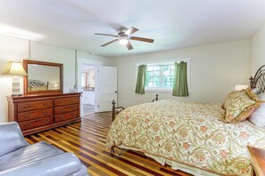 Quarto com cama, cômoda, cadeira, ventilador de teto e janela com cortinas.
