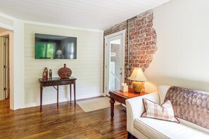 Uma sala de estar com piso de madeira, um sofá branco, uma mesa com abajur e uma televisão fixada na parede.