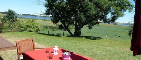 Un espacio de comedor al aire libre con una mesa dispuesta para dos, un árbol grande al fondo y vistas a una masa de agua.