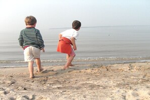 Dos niños jugando en una playa arenosa, con agua tranquila al fondo.