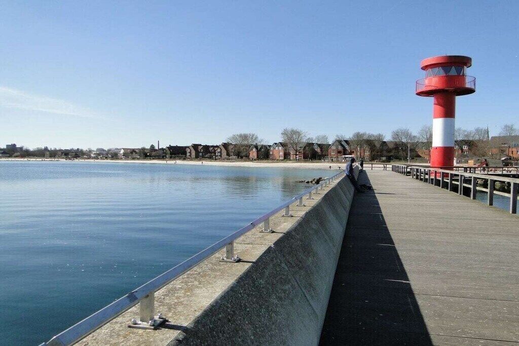 A waterfront promenade with a red and white lighthouse, a concrete barrier, and a row of buildings in the background.