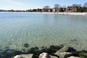 A clear lake with visible rocks at the shore, a sandy beach, and buildings in the background. - Above the Kattsund || Eckernförde (Eckernförde)