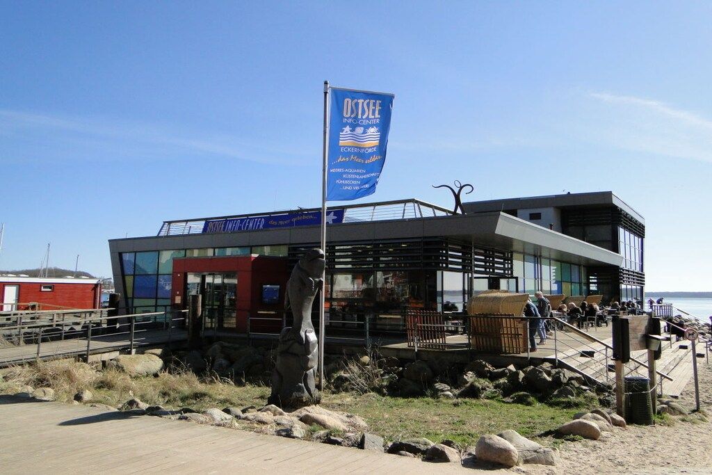 Un bâtiment moderne doté d’une façade en verre, d’un mât sur lequel flotte un drapeau bleu, et d’une promenade en bois menant à l’entrée.