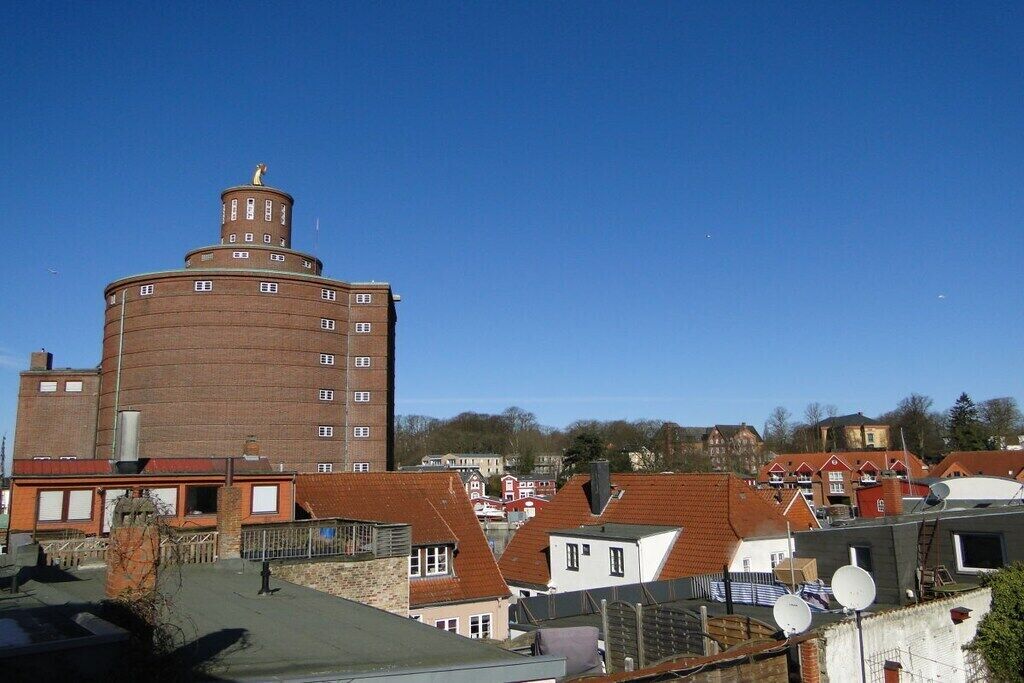 En stor murstensbygning med et cylindrisk tårn og en statue øverst, omgivet af boliger med røde tage.