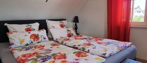 A double bed with floral bedding, a black headboard, a bedside lamp, and a window with red curtains.