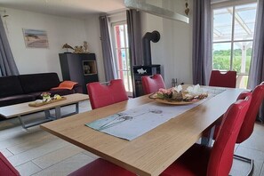 Ein modernes Wohnzimmer mit Esstisch, roten Stühlen, einer Couch, einem Kamin und Blick durch große Fenster.