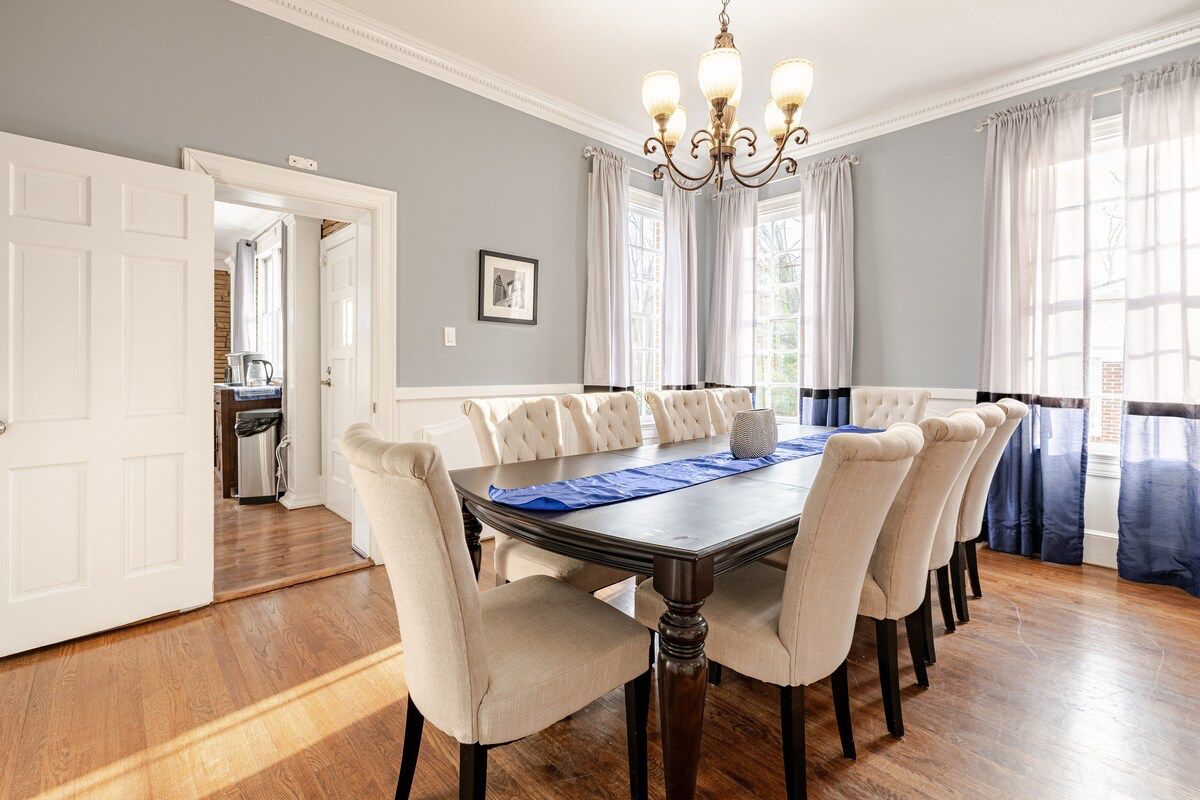A dining room with a large table, six chairs, a chandelier, and a view of a kitchen through an open door.