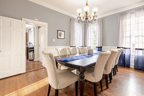 Une salle à manger avec une grande table, six chaises, un lustre et une vue sur la cuisine par une porte ouverte.