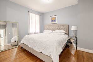 A bedroom with a bed, a mirror, a bedside table, and a window with curtains. - 6BR Fayetteville Retreat | Pool, Hot Tub & Pets (Fayetteville)