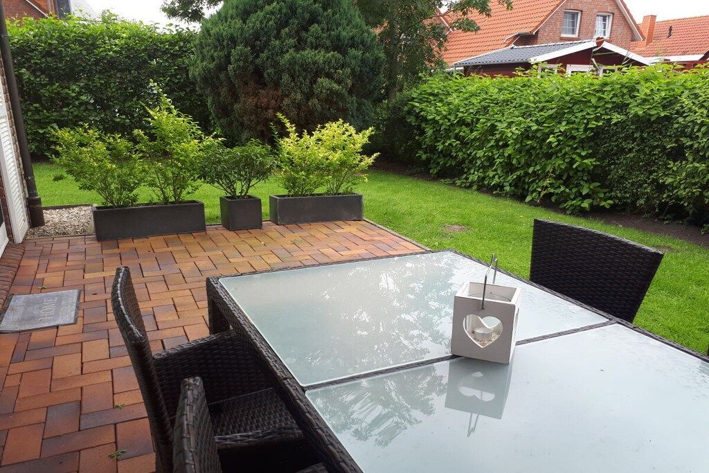 Un patio exterior con mesa y sillas de vidrio, rodeado de follaje y un piso de ladrillo.
