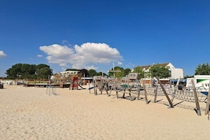 Une plage de sable bordée d’une rangée de chaises longues en bois et d’un filet de volley-ball, un petit kiosque et, au fond, un bâtiment à la façade blanche.