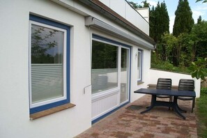 Ein modernes Gebäude mit großen Fenstern, einer Terrasse mit Tisch und Stühlen sowie einem gepflasterten Weg.