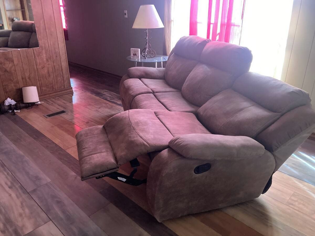 A reclining sofa in a living room with a wooden floor, a small table with a lamp, and a window with curtains.