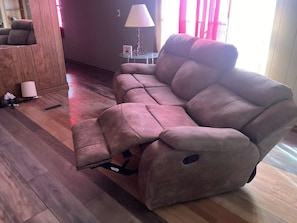 A reclining sofa in a living room with a wooden floor, a small table with a lamp, and a window with curtains.