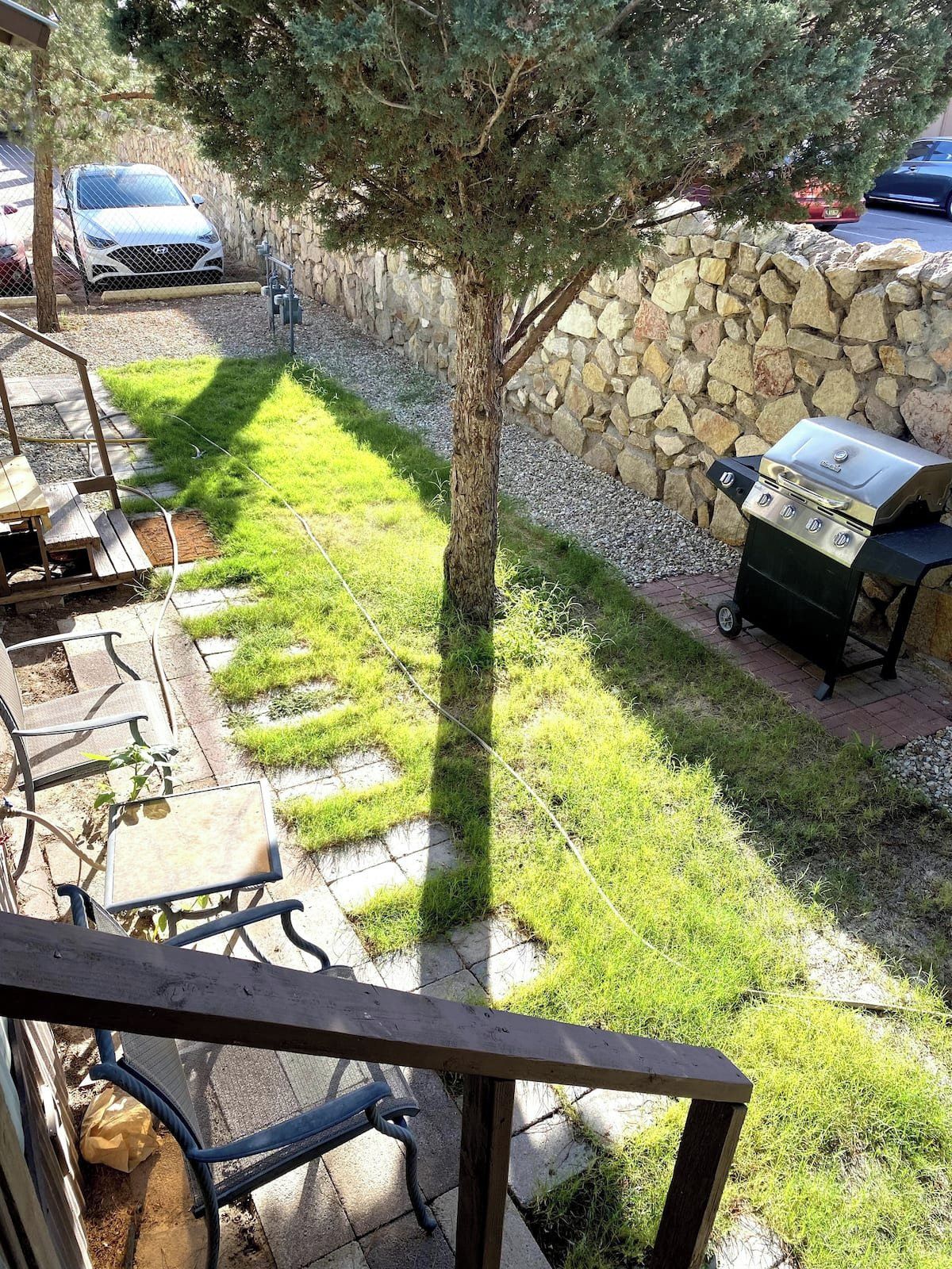 Un jardin avec un mur en pierre, un barbecue et une voiture garée au fond.