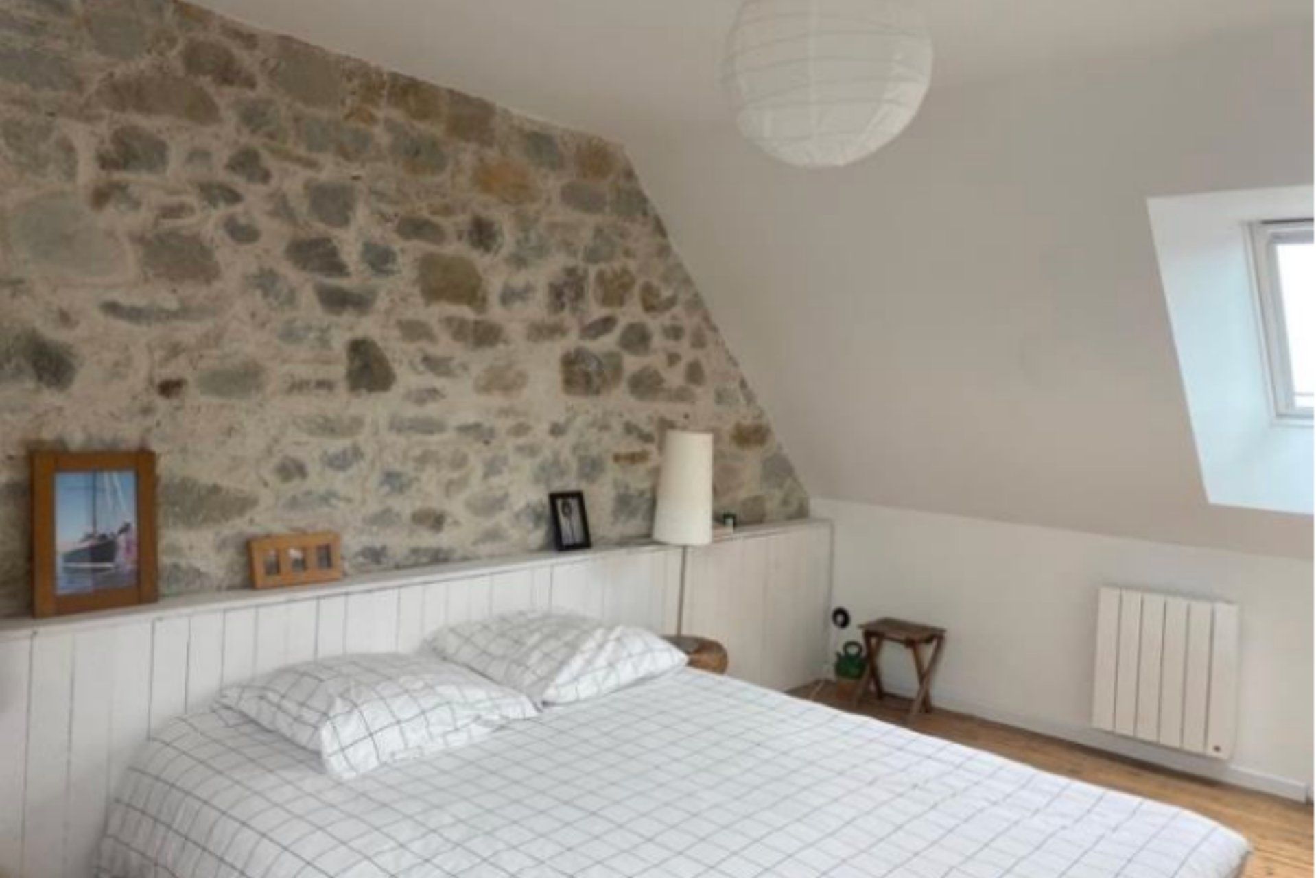 A bedroom with a stone wall, a bed with a white quilt, a bedside table with a lamp, and a window.